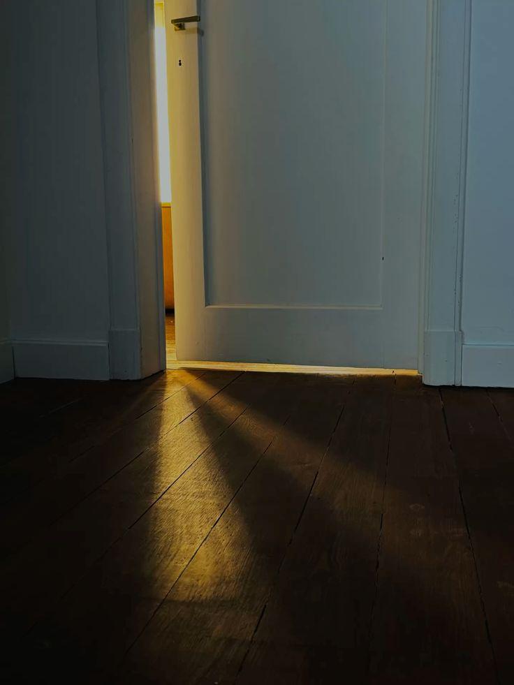 Puerta entreabierta en una habitación en penumbra; por la rendija entra luz cálida que se proyecta sobre el suelo de madera